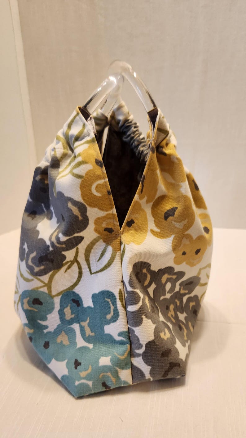 May include: A floral print tote bag with clear acrylic handles. The fabric features a pattern of yellow, blue, and gray flowers on a white background. The interior is a dark brown color.