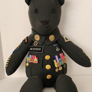 Op de afbeelding: Een knuffelbeer gekleed in een donkergroen militair uniform met gouden knopen, emblemen en medailles. Het naamplaatje luidt "MC KITRICK". De beer heeft zwarte knoopogen en een zwarte neus. De beer zit.