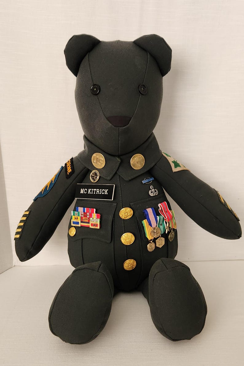 Op de afbeelding: Een knuffelbeer gekleed in een donkergroen militair uniform met gouden knopen, emblemen en medailles. Het naamplaatje luidt "MC KITRICK". De beer heeft zwarte knoopogen en een zwarte neus. De beer zit.