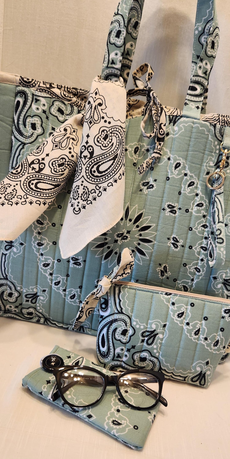 May include: A quilted tote bag, a matching zippered pouch, and a small rectangular case in a light sage green fabric featuring a black and white paisley bandana print. A white bandana with a black paisley pattern is tied to the tote handle.