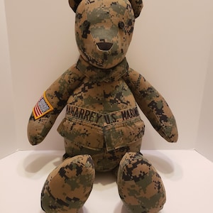 Op de afbeelding: Een handgemaakte teddybeer in een camouflagepatroon. De beer heeft knoopogen en een gestikte neus. Hij draagt een uniform met een Amerikaanse vlagpatch en de tekst "AHARREY U.S. MARINE".