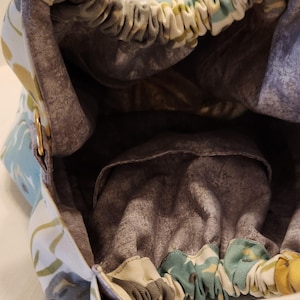 May include: An open view of a fabric handbag with a floral pattern in shades of blue, yellow, and green. The interior is a muted gray with a small pocket. The bag has a metal handle and a drawstring closure.