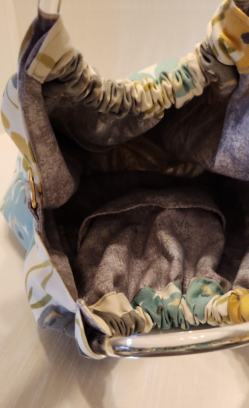 May include: An open view of a fabric handbag with a floral pattern in shades of blue, yellow, and green. The interior is a muted gray with a small pocket. The bag has a metal handle and a drawstring closure.