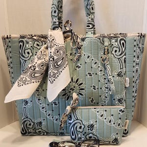 May include: A quilted tote bag, a matching zippered pouch, and a small rectangular case in a light sage green fabric with a black and white paisley bandana print. The tote features two handles, a tied bandana accent, and a small metal key ring.