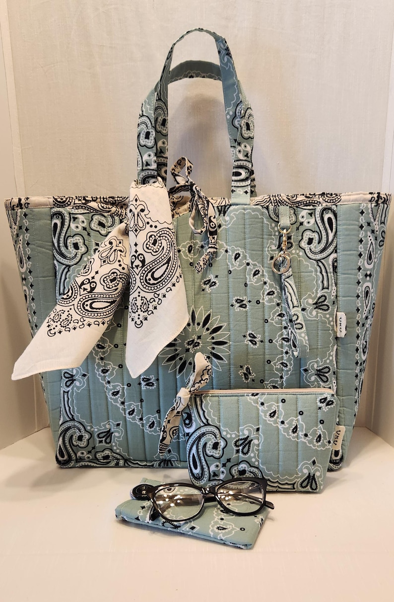 May include: A quilted tote bag, a matching zippered pouch, and a small rectangular case in a light sage green fabric with a black and white paisley bandana print. The tote features two handles, a tied bandana accent, and a small metal key ring.