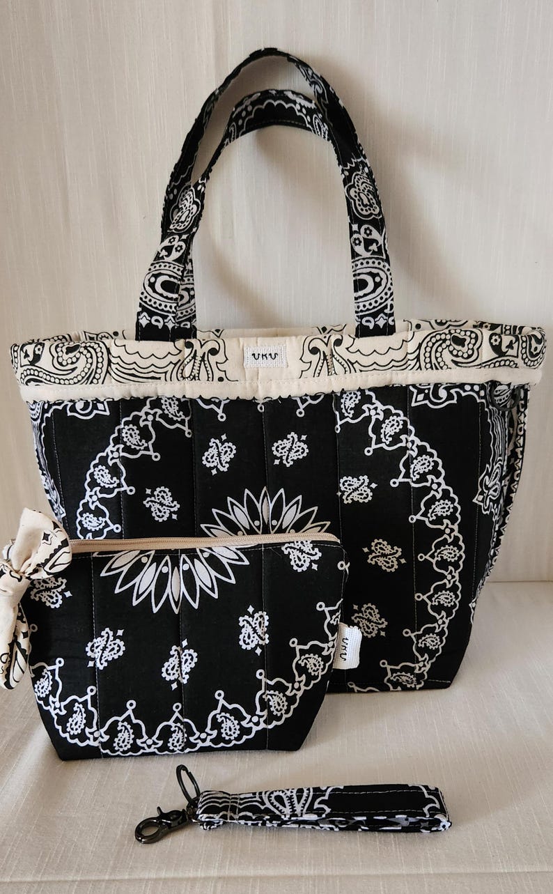 May include: A black and white quilted tote bag, a matching zippered pouch with a side bow, and a wristlet strap. Each item features a repeating paisley bandana pattern. The tote and pouch have a small white label with the text UKU.