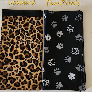 May include: Two rectangular fabric pieces displayed side by side. The left piece features a brown and black leopard print pattern with a solid black top border. The right piece is black with a repeating pattern of grey and white paw prints.