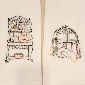 May include: Two white kitchen towels with embroidered designs. One features a decorative shelf with kitchen items, the other a pot rack with hanging utensils. The embroidery uses black, red, yellow, and blue thread.