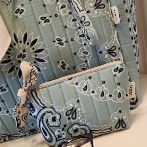 May include: A quilted sage green tote bag and matching zippered pouch featuring a black and white paisley bandana print. The set includes a small fabric knot detail on the pouch and a gold-toned metal clip. Black-rimmed glasses rest in front.