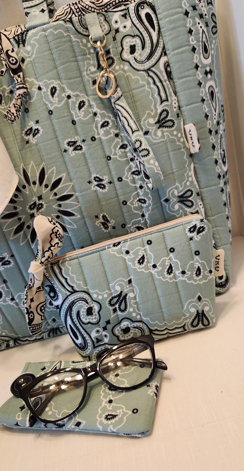 May include: A quilted sage green tote bag and matching zippered pouch featuring a black and white paisley bandana print. The set includes a small fabric knot detail on the pouch and a gold-toned metal clip. Black-rimmed glasses rest in front.