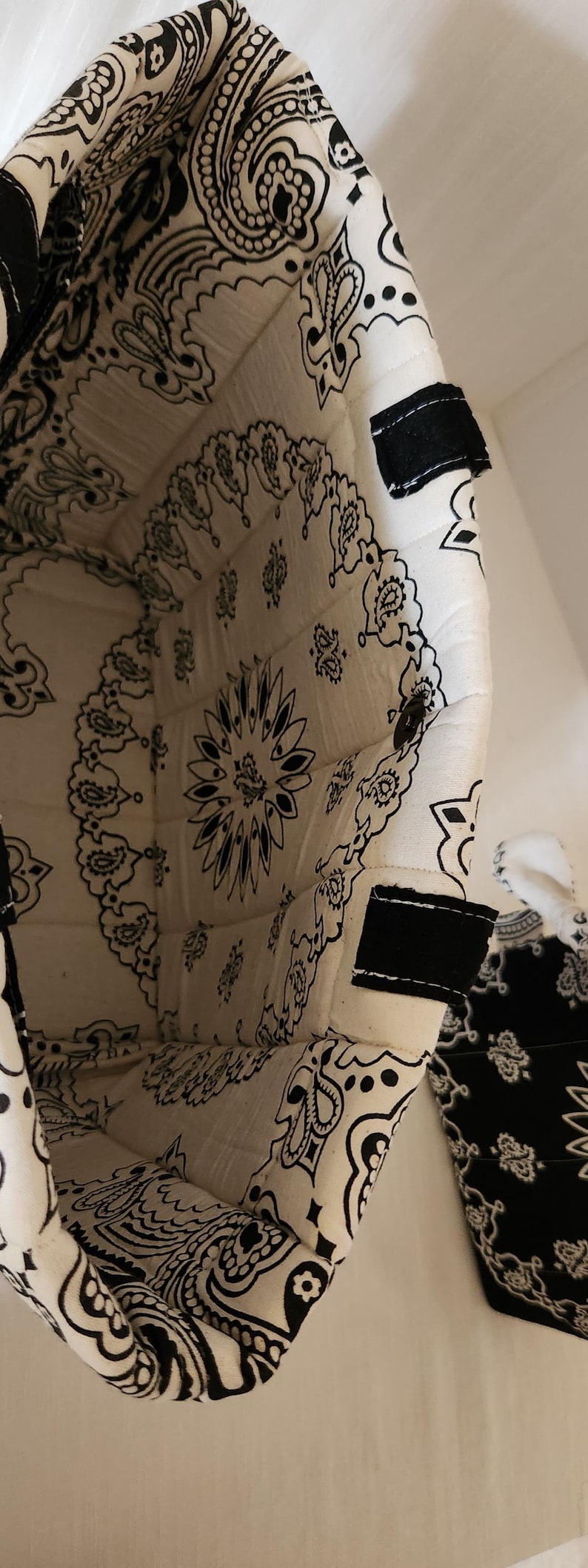 May include: A quilted fabric basket featuring a black and white paisley bandana print. The interior and exterior are lined with the same patterned textile, and the rim includes black fabric tabs. The structure is soft, flexible, and cylindrical.