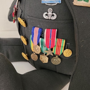 Op de afbeelding: Een knuffelbeer gekleed in een donkergroen militair uniform, versierd met diverse medailles, linten en insignes. Het uniform heeft gouden knopen, een patch met groene bladeren en een parachute-embleem. De arm van de beer is zichtbaar aan de linkerkant.