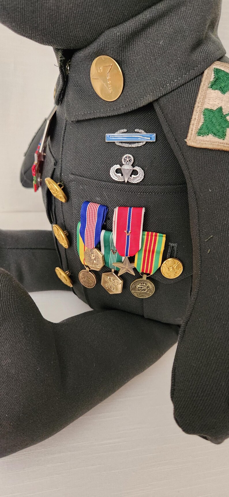 Op de afbeelding: Een knuffelbeer gekleed in een donkergroen militair uniform, versierd met diverse medailles, linten en insignes. Het uniform heeft gouden knopen, een patch met groene bladeren en een parachute-embleem. De arm van de beer is zichtbaar aan de linkerkant.