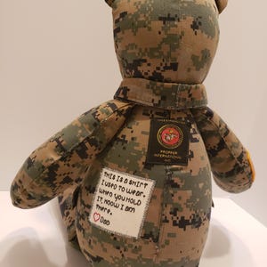 Op de afbeelding: Een handgemaakte teddybeer gemaakt van camouflage stof, met een kraag en patches. E&eacute;n patch luidt "THIS IS A SHIRT I USED TO WEAR. WHEN YOU HOLD IT, KNOW I AM THERE. Dad" met een hart. Een andere patch luidt "PROPPER INTERNATIONAL INC."