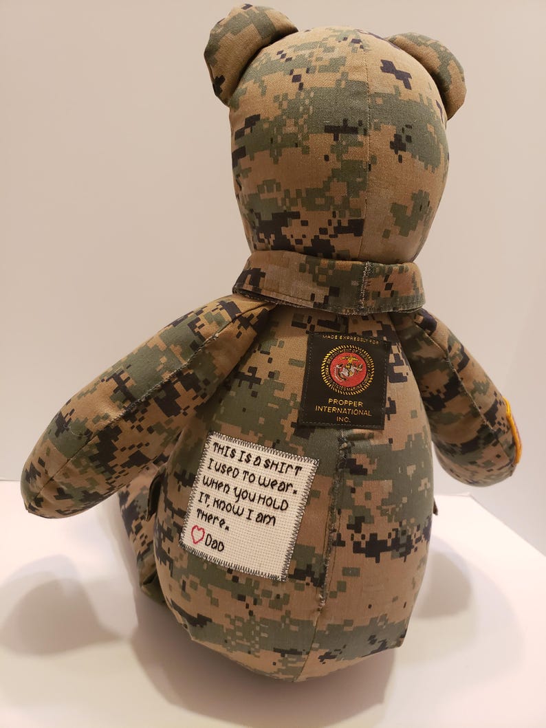 Op de afbeelding: Een handgemaakte teddybeer gemaakt van camouflage stof, met een kraag en patches. E&eacute;n patch luidt "THIS IS A SHIRT I USED TO WEAR. WHEN YOU HOLD IT, KNOW I AM THERE. Dad" met een hart. Een andere patch luidt "PROPPER INTERNATIONAL INC."