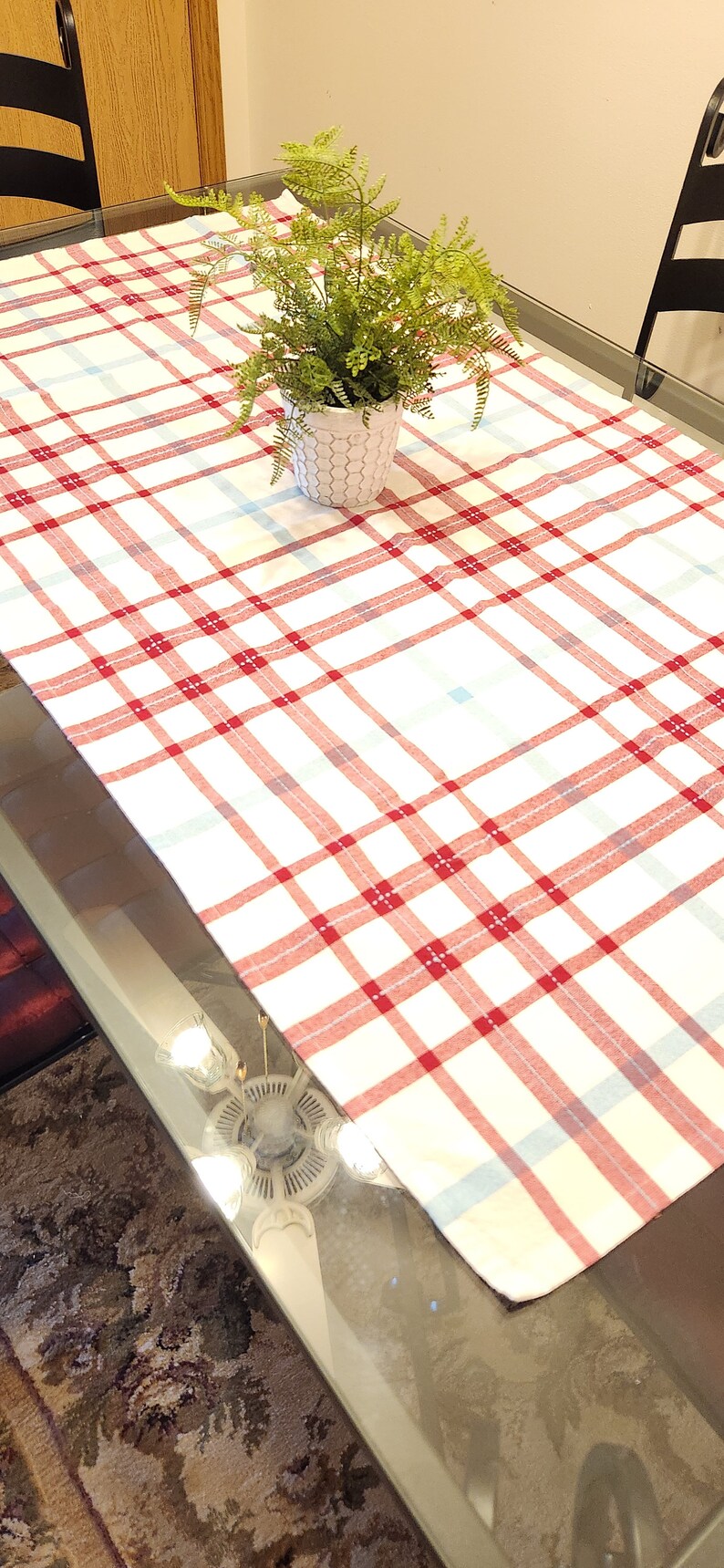 K&ouml;nnte beinhalten: Eine wei&szlig;e Tischdecke mit rotem und blauem Karomuster auf einem Glastisch mit einer Topfpflanze in der Mitte.