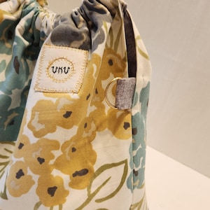 May include: A floral tote bag with a clear acrylic handle. The bag features a cream-colored background with yellow, blue, and green floral patterns. A small patch with embroidered text is visible on the front. The bag has a gray fabric accent and a gold-colored ring.