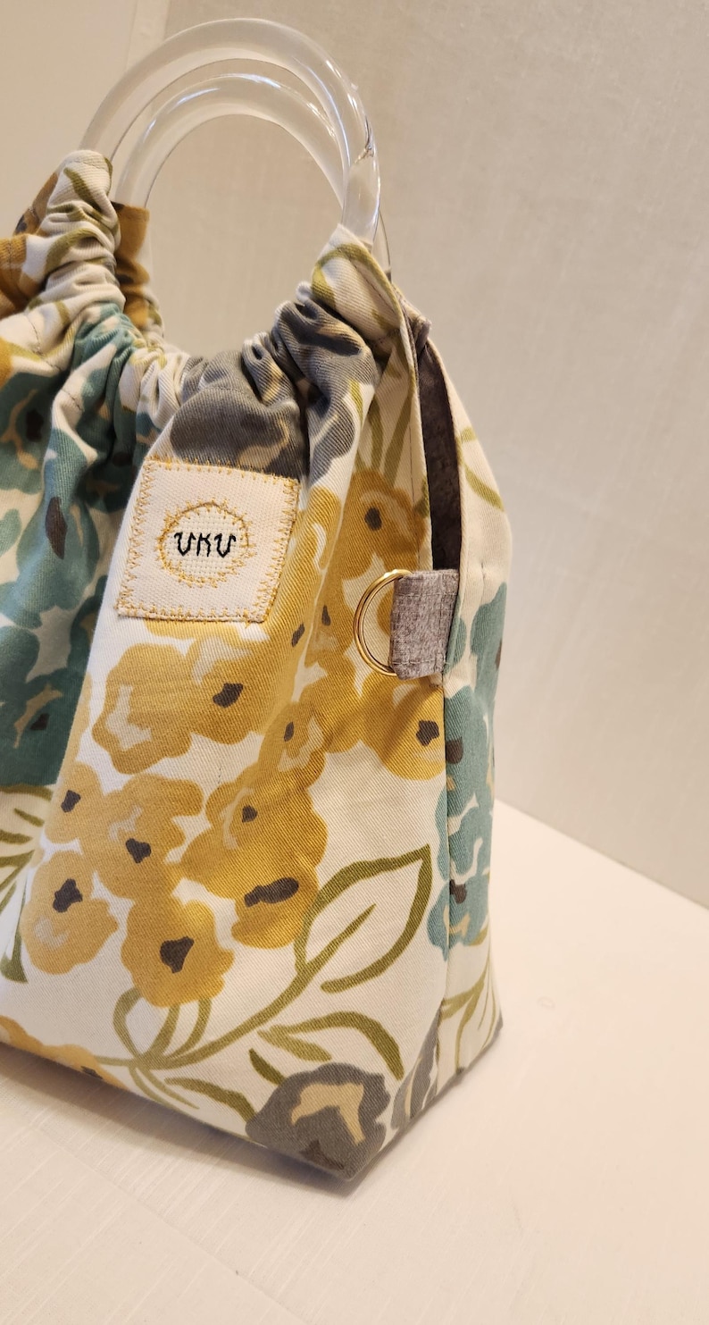 May include: A floral tote bag with a clear acrylic handle. The bag features a cream-colored background with yellow, blue, and green floral patterns. A small patch with embroidered text is visible on the front. The bag has a gray fabric accent and a gold-colored ring.