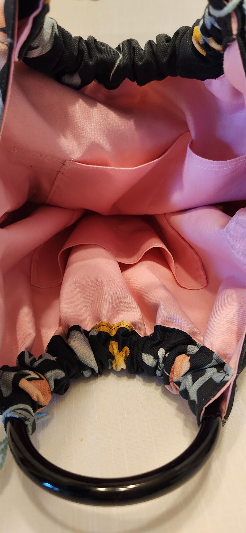 May include: A handmade tote bag with a black floral pattern on the exterior and a light pink interior. The bag has a black handle and several interior pockets. The floral pattern includes yellow, gray, and peach-colored flowers.