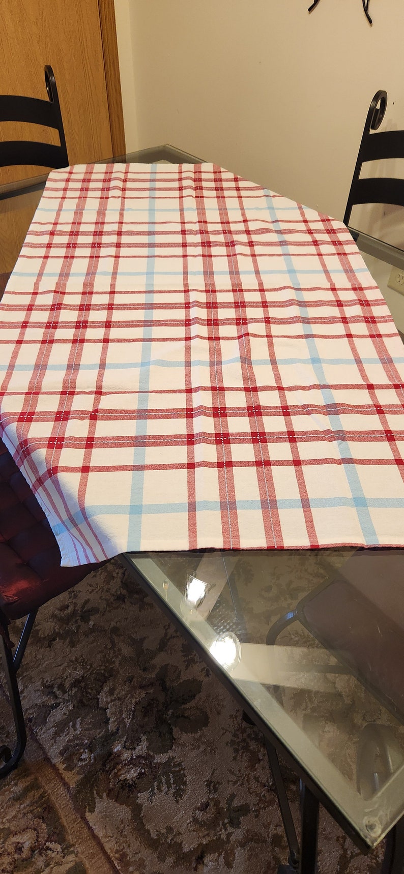 K&ouml;nnte beinhalten: Eine rot-blaue karierte Tischdecke auf einem Glastisch mit zwei schwarzen St&uuml;hlen. Die Tischdecke ist in der Mitte gefaltet.