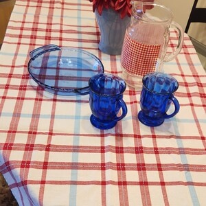 K&ouml;nnte beinhalten: Eine rot-wei&szlig; karierte Tischdecke mit zwei blauen Glasbechern und einer blauen Glas-Tortenplatte. Eine Vase mit roten Blumen steht auf dem Tisch.