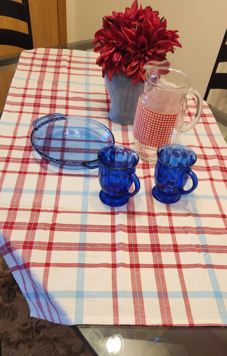K&ouml;nnte beinhalten: Eine rot-wei&szlig; karierte Tischdecke mit zwei blauen Glasbechern und einer blauen Glas-Tortenplatte. Eine Vase mit roten Blumen steht auf dem Tisch.