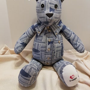 May include: A blue and white patterned teddy bear wearing a shirt with buttons. The bear is sitting on a white fabric surface. The bear's right foot has a white patch with the word "Grandpa" embroidered in red.
