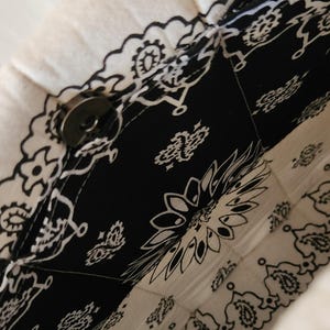 May include: A quilted fabric bag featuring a black and white bandana print with intricate paisley and floral patterns. The bag is constructed with stitched panels and includes a circular metallic magnetic snap closure at the top opening.