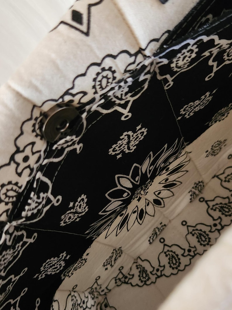 May include: A quilted fabric bag featuring a black and white bandana print with intricate paisley and floral patterns. The bag is constructed with stitched panels and includes a circular metallic magnetic snap closure at the top opening.