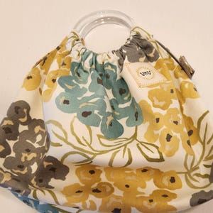 May include: A floral print handbag with a clear, rounded handle. The bag features a cream-colored background with a pattern of yellow, teal, and gray flowers. A small tag with the letters "umu" is attached to the bag.
