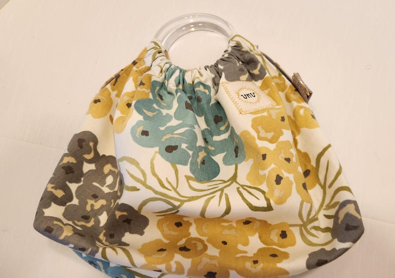 May include: A floral print handbag with a clear, rounded handle. The bag features a cream-colored background with a pattern of yellow, teal, and gray flowers. A small tag with the letters "umu" is attached to the bag.