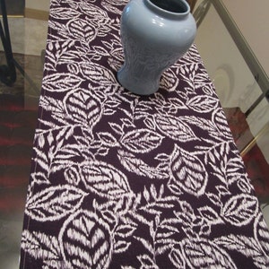 May include: A purple table runner with a white floral pattern and a fringed edge. A light blue ceramic vase is on top of the runner.