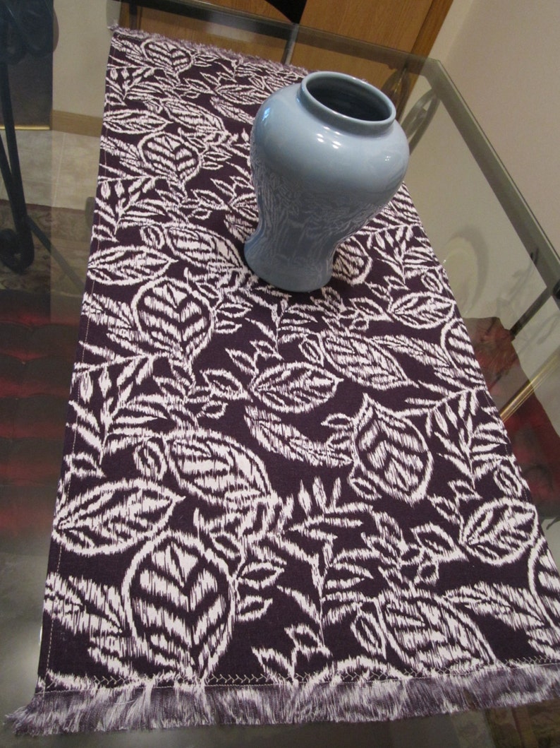 May include: A purple table runner with a white floral pattern and a fringed edge. A light blue ceramic vase is on top of the runner.
