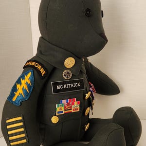 Op de afbeelding: Een donkergroene teddybeer met een militair uniform. Het uniform heeft patches met "AIRBORNE" en "MC KITRICK". De beer heeft gouden knopen en diverse medailles.