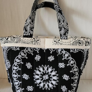 May include: A quilted tote bag featuring a black and white bandana print with a central floral motif. The bag has two matching fabric handles and a cream-colored fabric trim along the top edge, showcasing a structured, vertical stitched design.