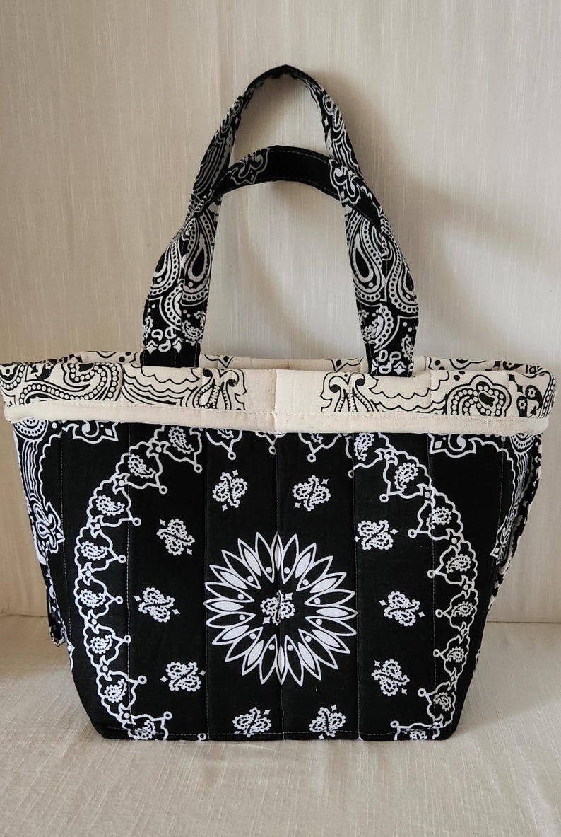 May include: A quilted tote bag featuring a black and white bandana print with a central floral motif. The bag has two matching fabric handles and a cream-colored fabric trim along the top edge, showcasing a structured, vertical stitched design.
