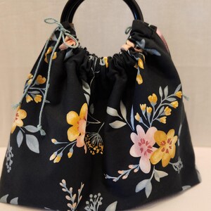 May include: A black drawstring bag with a floral pattern featuring yellow and pink flowers, and gray leaves. The bag has a black handle and light blue drawstrings.
