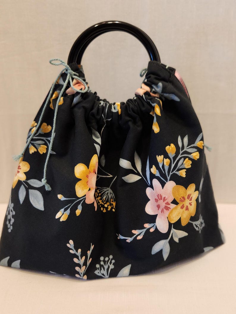 May include: A black drawstring bag with a floral pattern featuring yellow and pink flowers, and gray leaves. The bag has a black handle and light blue drawstrings.