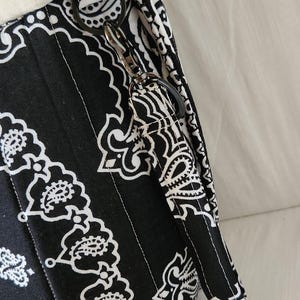 May include: A black and white fabric bag featuring a repeating paisley bandana pattern. The bag includes a cream-colored fabric trim at the top edge and a matching fabric wrist strap attached to a black metal D-ring with a swivel lobster clasp.