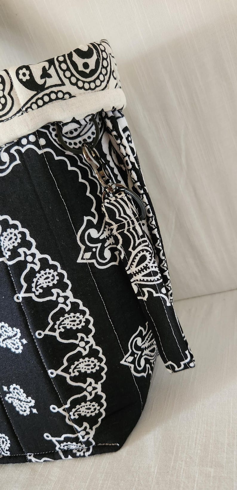 May include: A black and white fabric bag featuring a repeating paisley bandana pattern. The bag includes a cream-colored fabric trim at the top edge and a matching fabric wrist strap attached to a black metal D-ring with a swivel lobster clasp.