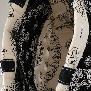 May include: A quilted tote bag featuring a black and white paisley bandana print. The interior is lined with the same pattern and includes a side pocket with a metal snap closure. A small white rectangular tag with black text is sewn on the rim.