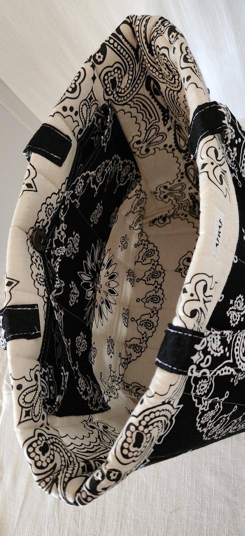 May include: A quilted tote bag featuring a black and white paisley bandana print. The interior is lined with the same pattern and includes a side pocket with a metal snap closure. A small white rectangular tag with black text is sewn on the rim.