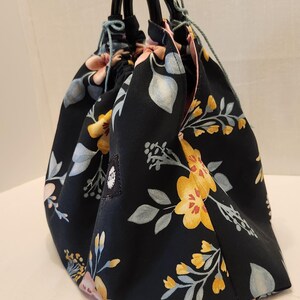 May include: A black floral print bag with a drawstring closure and black handles. The bag features a pattern of yellow, pink, and blue flowers and leaves. The bag is made of fabric and has a rounded shape.