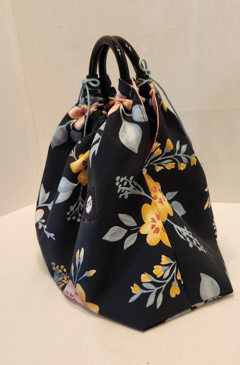 May include: A black floral print bag with a drawstring closure and black handles. The bag features a pattern of yellow, pink, and blue flowers and leaves. The bag is made of fabric and has a rounded shape.