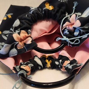 May include: A handmade drawstring bag with a floral pattern in navy blue, peach, and white. The bag has black circular handles and a light pink interior. Light blue drawstrings are visible.