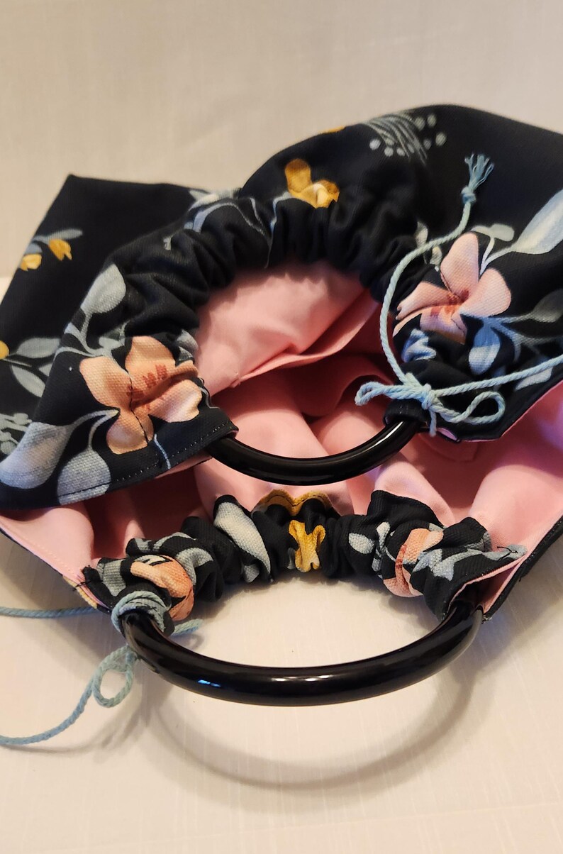 May include: A handmade drawstring bag with a floral pattern in navy blue, peach, and white. The bag has black circular handles and a light pink interior. Light blue drawstrings are visible.