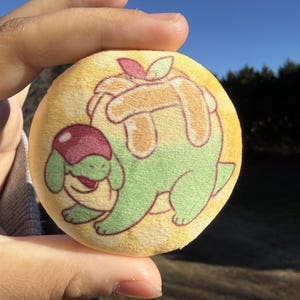May include: A round, yellow plushie featuring a green cartoon character with a red and green fruit design. The plushie is held in a hand against a backdrop of a blue sky and trees.