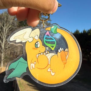 May include: A clear acrylic keychain featuring a cartoon dragon in shades of yellow, orange, and green. The dragon is curled up with a rainbow-colored gem. The keychain has a gold-colored clasp.