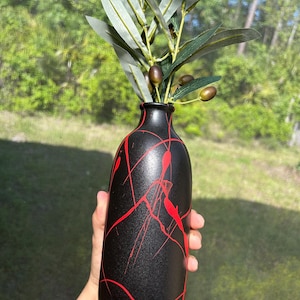 May include: A black vase with red paint splatter design, holding olive branches with green leaves and olives. The vase has a narrow neck and a rounded body. The background is a blurred view of green foliage.