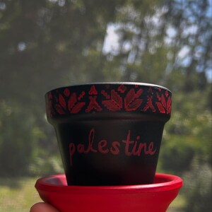 May include: A black flower pot with red floral designs and the word "palestine" in red script. The pot sits on a red saucer. The background is blurred green foliage.
